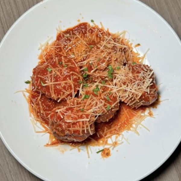 Four meatballs covered in red sauce, shredded Parmesan cheese, and green herbs on a white plate.