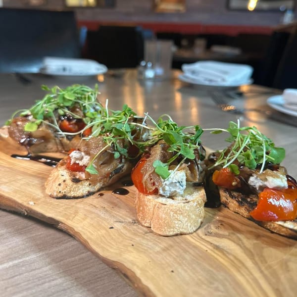 Four bruschetta pieces with caramelized onions, tomatoes, cheese, and microgreens on a wooden board.