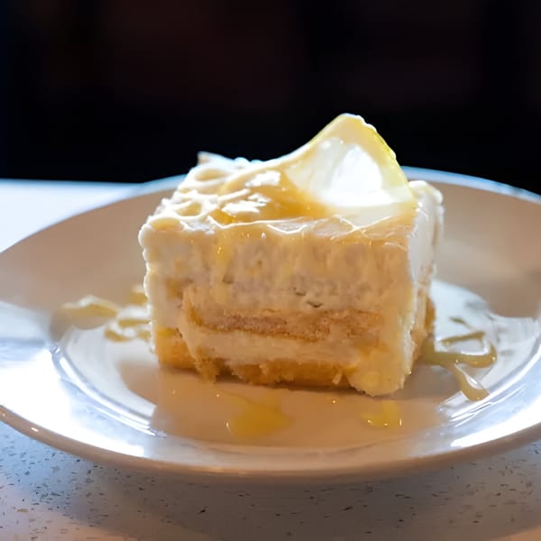 A slice of lemon cake with white frosting and a lemon slice on a white plate.