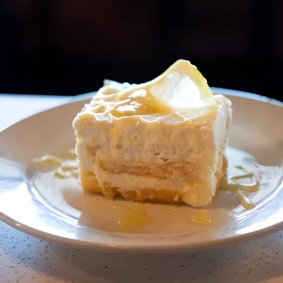 A slice of lemon cake with white frosting and a lemon slice on a white plate.