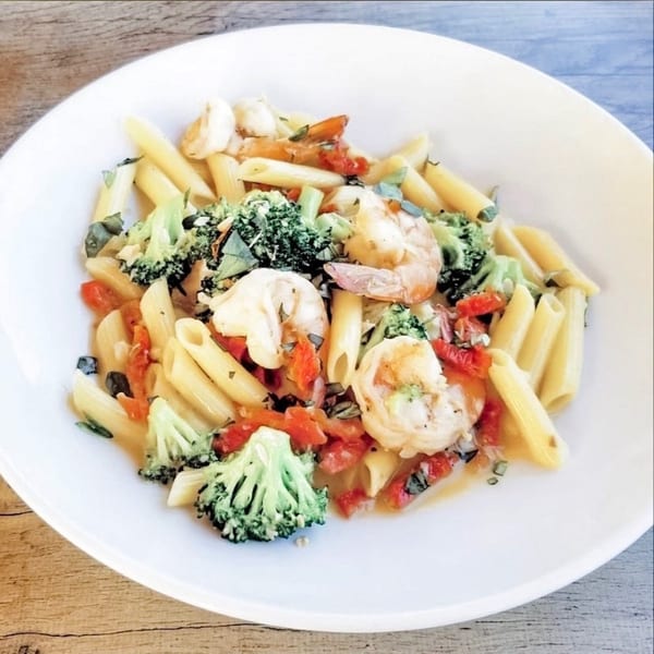 Penne pasta with shrimp, broccoli, and diced tomatoes in a light sauce, served in a white bowl.